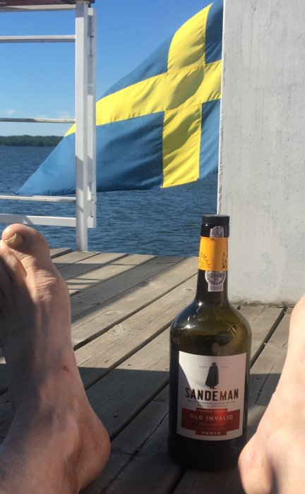 Person med fötterna upp, flaskan portvin i förgrunden, svensk flagga och sjö i bakgrunden.