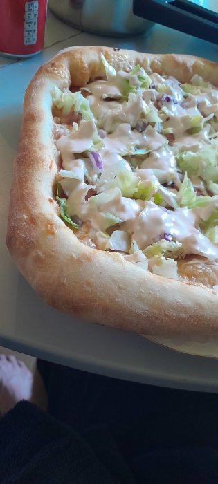 Hemlagad pizza med tjock botten, sallad och vit sås på ett köksbord.