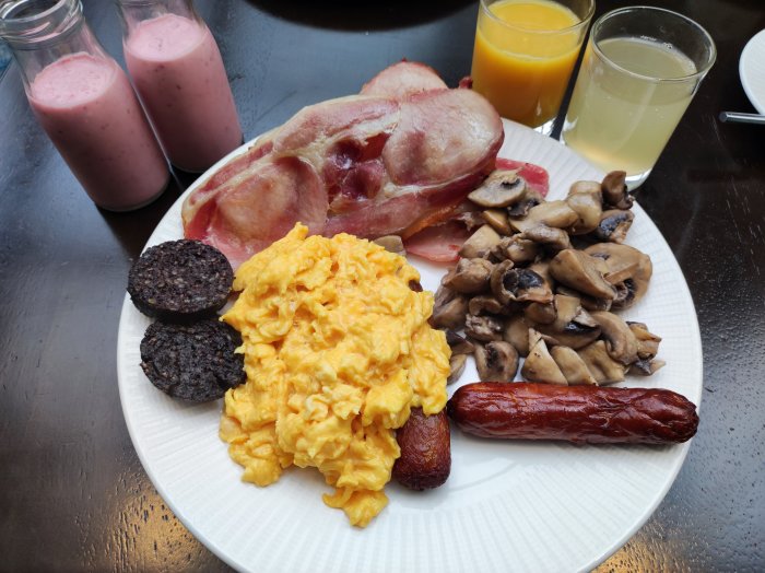 Frukosttallrik med äggröra, bacon, korv, blodpudding, champinjoner, smoothie och juice.