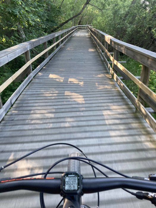Cyklistens perspektiv på en träbro i grönt landskap med cykelstyrets topp och cykeldator i förgrunden.