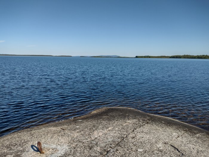 Klippstrand vid vatten i Lapplands skärgård med blå himmel och par solglasögon på marken.