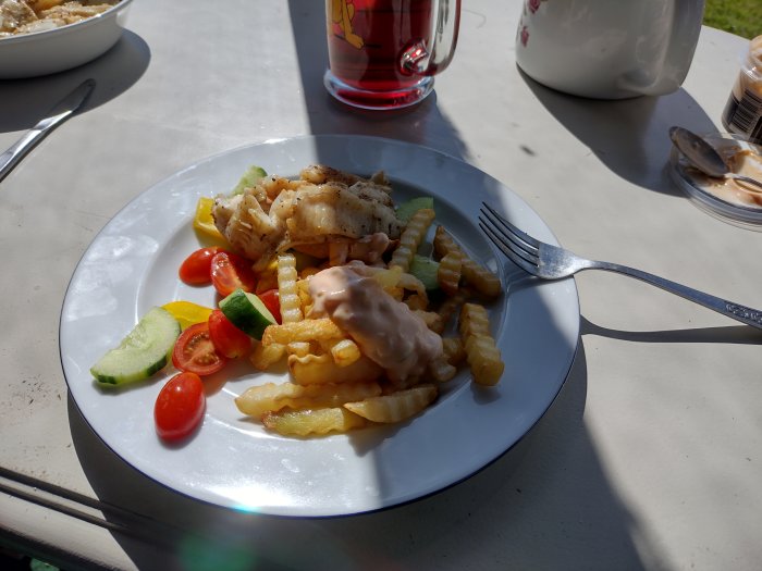 Tallrik med fisk, pommes frites, grönsaker och rörsås på ett bord utomhus i soligt väder.