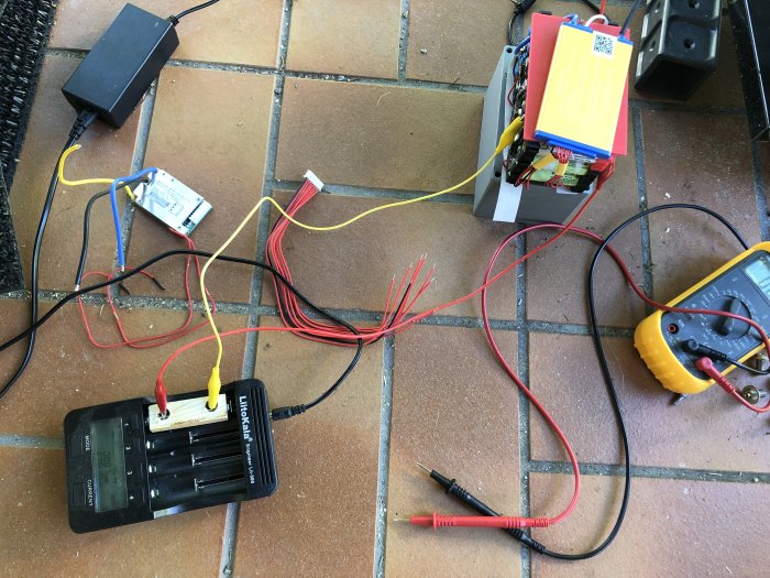 Adapter och batterier inkopplade till en laddare med multimeter på kakelgolv.