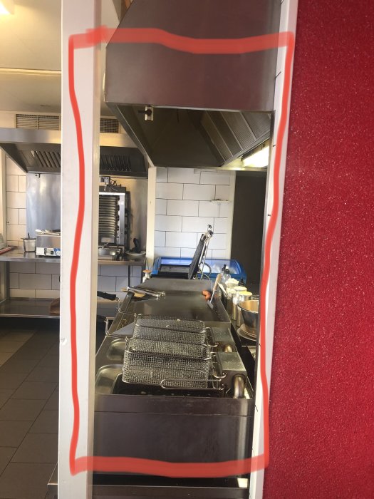 Vy genom fönster av restaurangkök med fritöser och utrustning, arbetsytor och hyllor i bakgrunden.