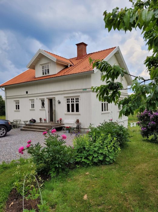 Trädgård med blommande buskar framför en vit trävilla med tegeltak på en sommardag.