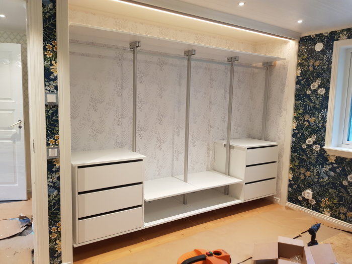 En nyinstallerad vit garderob med öppna moduler och lådor i ett rum med mönstrade tapeter och golv i renoveringsfas.
