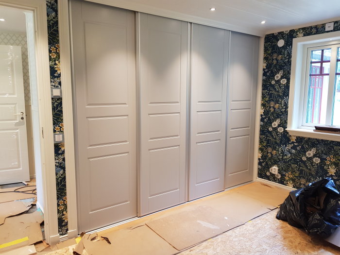 Inbyggda garderober med grå dörrar inne i ett rum med blommigt tapetserade väggar. Renovering pågår.