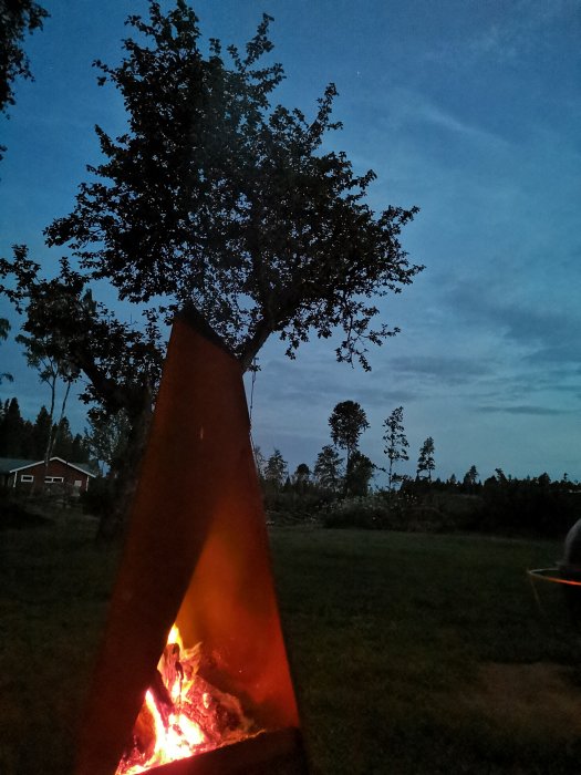 Kvällsbild av upplyst Tipi-liknande eldstad med levande eld på gräsmatta med träd i bakgrunden.