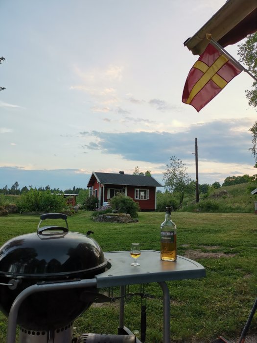 Utsikt över en trädgård med en flaska whisky och ett glas på ett bord, röd stuga och svensk flagga i bakgrunden.