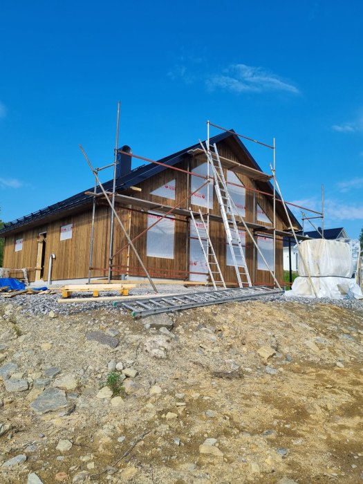 Halvfärdigt hus med en klar gavel, byggställning, och panelering under blå himmel.