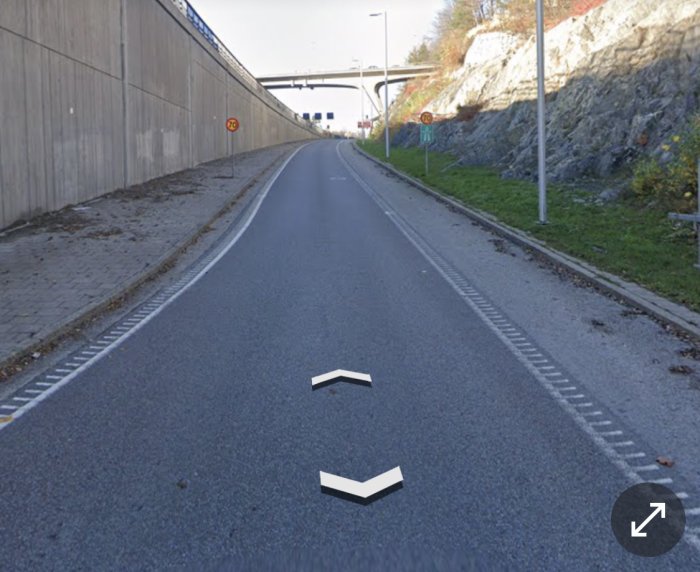 Påfartsramp med tom vägbana och vägmarkeringar, omgivet av betongvägg och klippa.