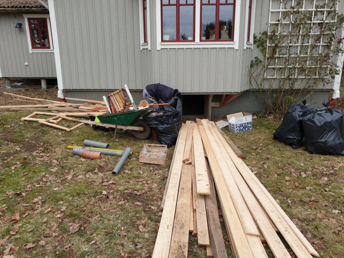 Virke och byggmaterial framför hus med skottkärra, plastpåsar och rör på gräsmatta.