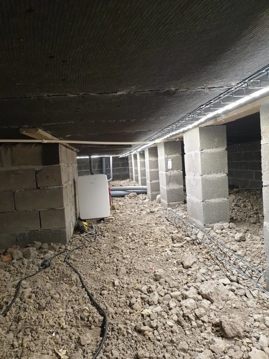 Ojämnt underlag och installationer under en husgrund med synliga betongpelare och isoleringsmaterial.