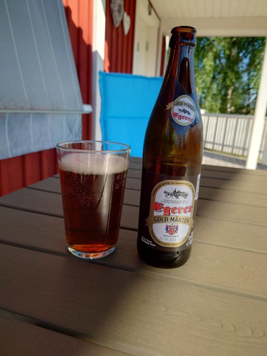 Flaska och glas med mörk öl på ett träbord utomhus, "Gold Märzen" märkt på flaskan.