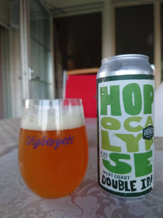 Ett glas med öl bredvid en burk Hopocalypse West Coast Double IPA på ett bord med röd stol i bakgrunden.