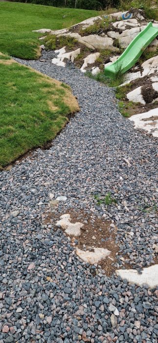 Grusgång med dräneringsstenar bredvid gräsmatta och plastlekrutschkana i en trädgård.