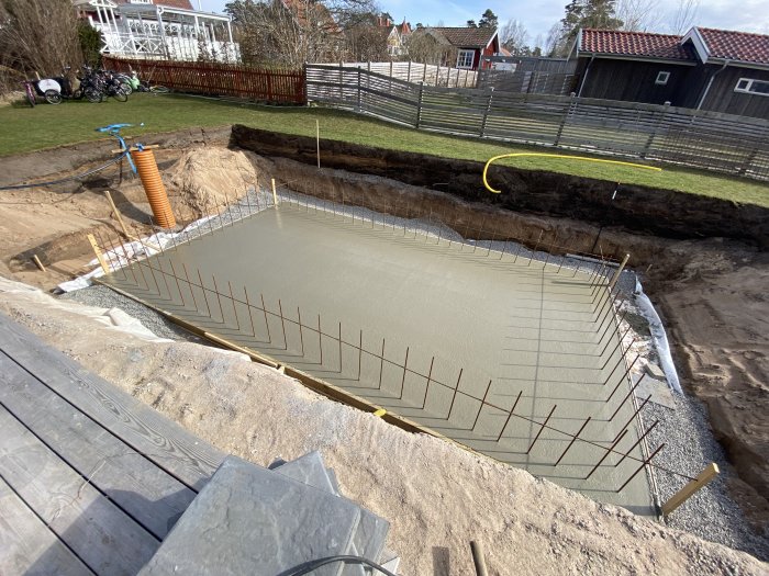 Nybyggd poolgrund i trädgård, med armeringsjärn och nylagt betong, omgiven av grävd jord.