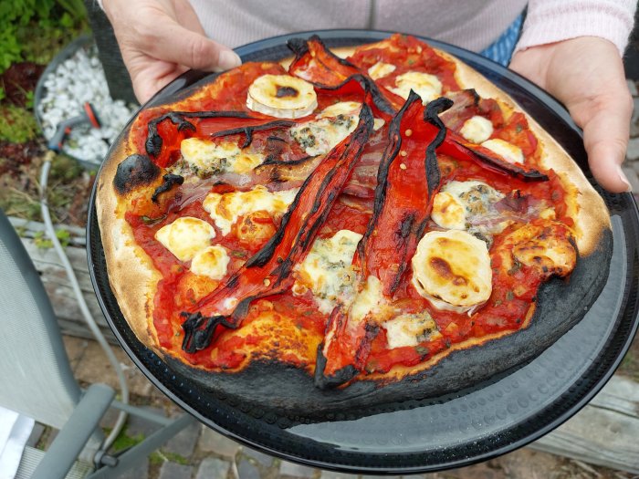 Hemgjord pizza med brända kanter, paprika och ost på en svart pizzaspade utomhus.