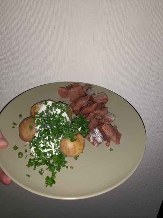 Ett fat med sill, kokt potatis, gräddfil och hackad gräslök presenterat mot en ljus bakgrund.