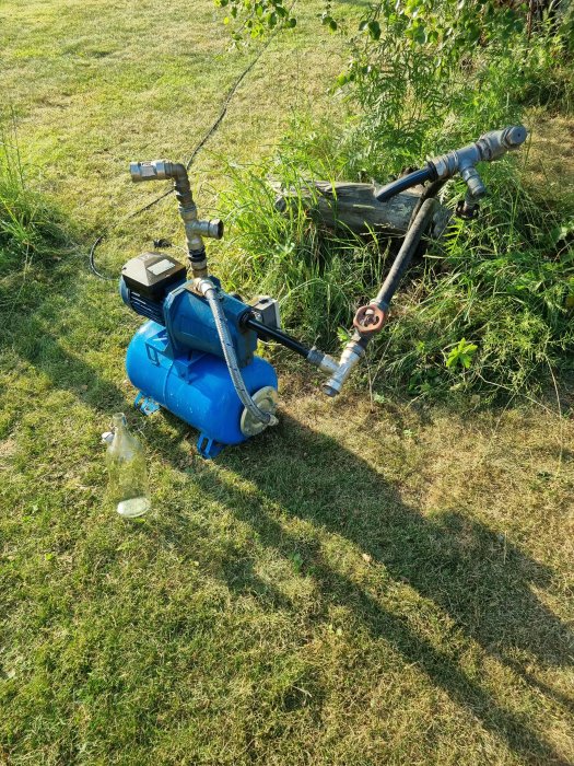 En blå vattenpump på gräsmatta med anslutna slangar och en tom glasflaska på marken.