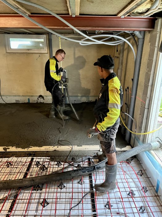 Två arbetare utför gjutningsarbete på en armerad betongplatta inomhus.