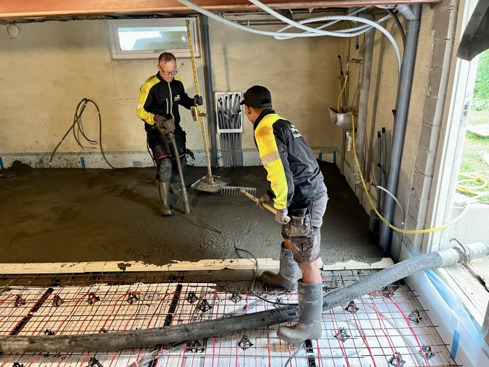Två arbetare jämnar ut nygjuten betong i ett byggnadsprojekt, en med spade och en med vibreringsverktyg.