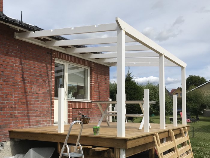 Delvis byggt trädäck i en trädgård med nyinstallerade vita reglar och läkter på en pergola-liknande konstruktion.