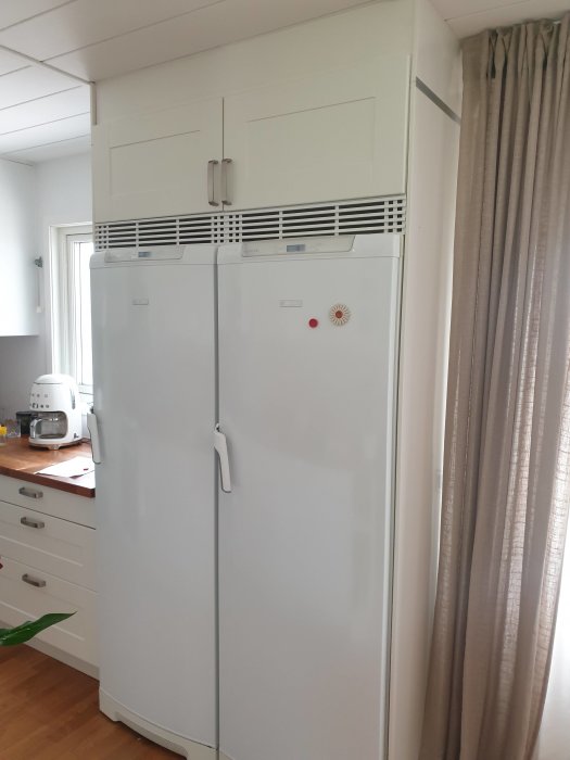 Ett vit kyl och frys par under skåp och galler med utrymme ovanpå i ett kök, vid sidan av ett fönster och gardiner.