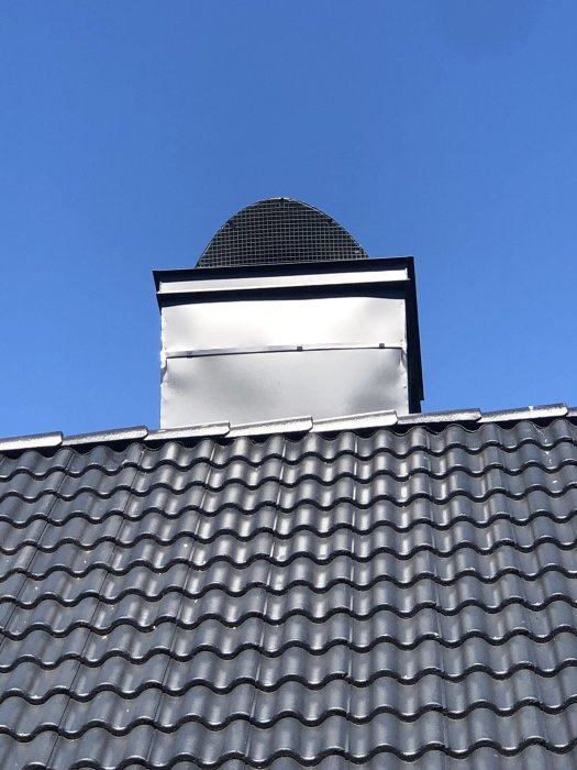 Inplåtad skorsten på ett svart tak med synliga veck och ojämnheter mot en klarblå himmel.