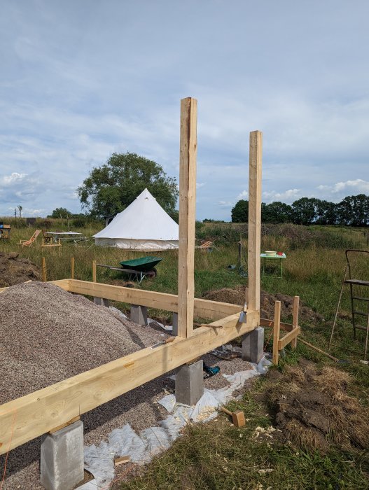 Grundläggning och stolpar av trä för pågående byggprojekt, med tält och byggmaterial i bakgrunden.