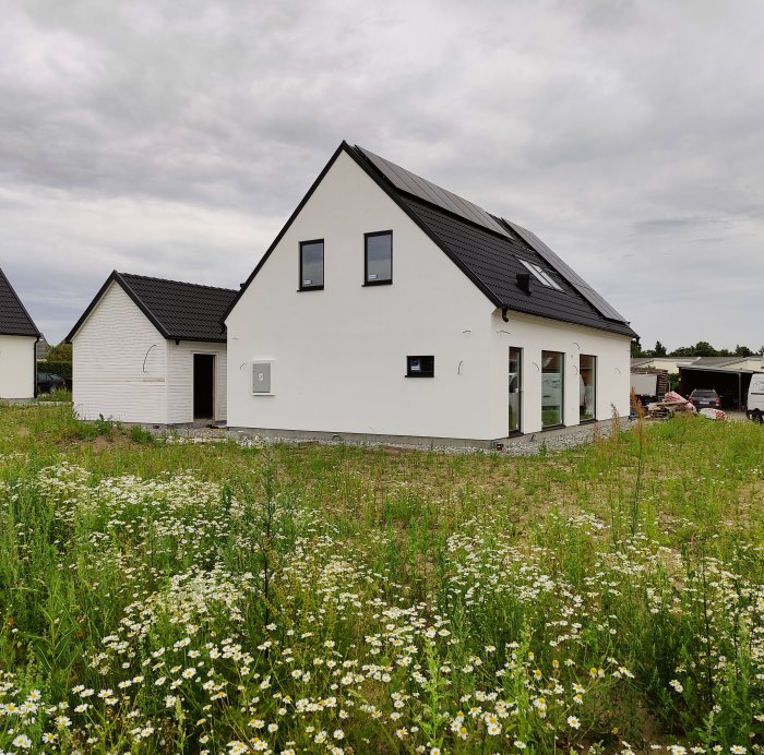 Nybyggt hus med solpaneler på taket och vit fasad, omgivet av en blommande äng.