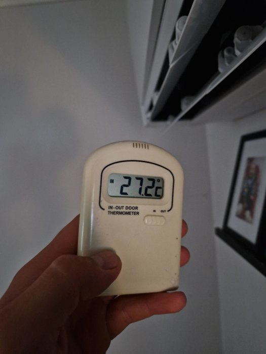 Hand håller en inomhus-utomhustermometer som visar 27.22 grader Celsius.