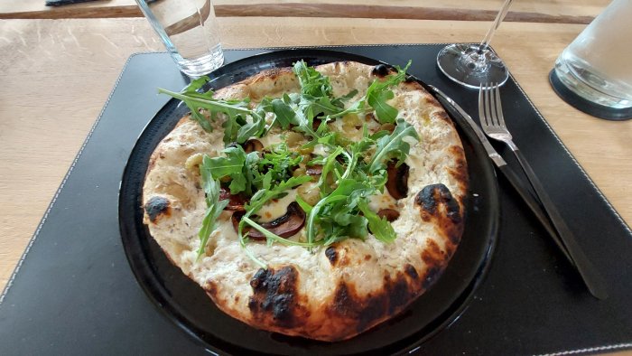 Hembakad pizza med välgräddad botten, toppad med ost och ruccola, på svart tallrik med bestick.