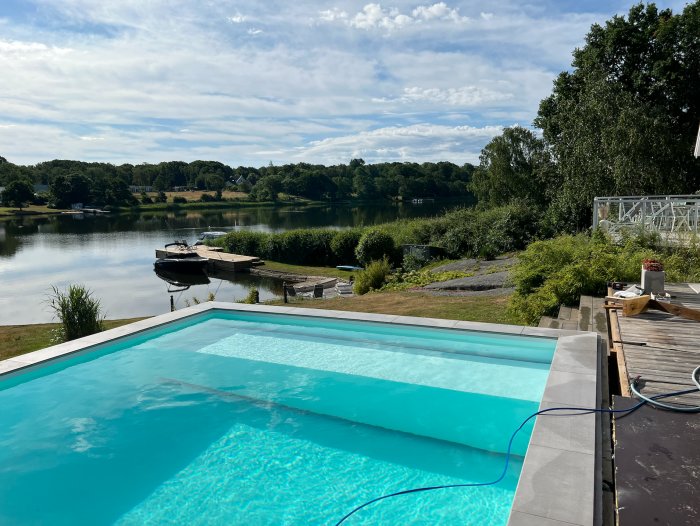 Utomhuspool med klart vatten intill en sjö med båt och brygga, omgiven av trädgård och himmel.