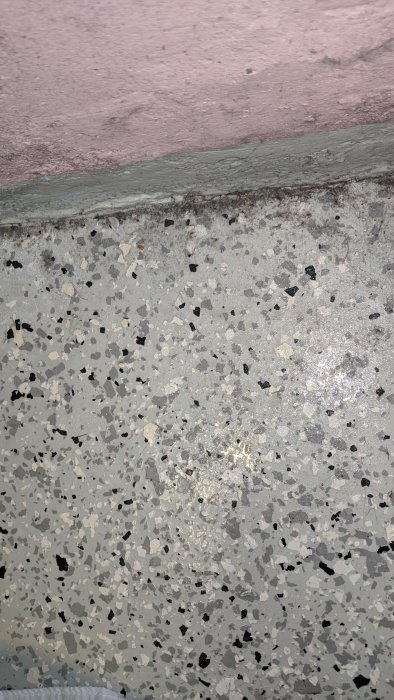 Fukt eller kondens på ett källargolv nära väggen, med synliga vattendroppar på ett terrazzoliknande golv.
