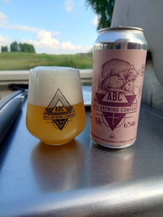 Ett glas med öl och en burk från APEX Brewing Company på en metallisk yta utomhus.