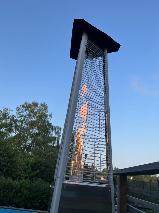 Utomhus gasvärmare med flammor bakom skyddsnät vid skymning.
