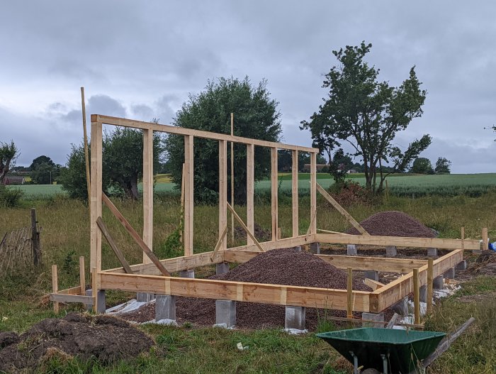 Nybyggd långsida av stomme med stolpar och hammarband i ett byggnadsprojekt, två grushögar i förgrunden.