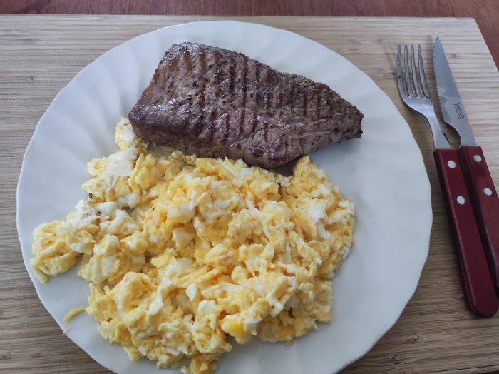 Grillad biff med stekränder och äggröra på en vit tallrik med gaffel och kniv vid sidan.