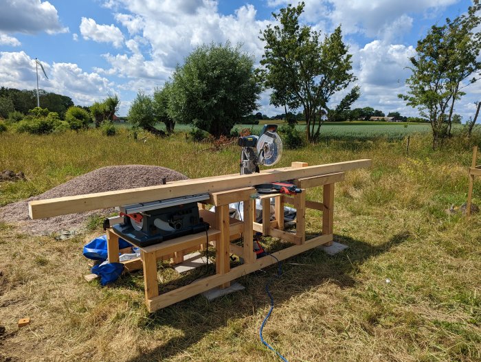 Ej helt färdigställd snickarbänk i trä med monterad kap- och klyvsåg utomhus på ett gräsbevuxet fält.