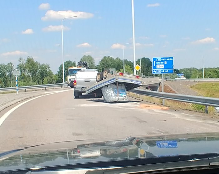 Vält släpvagn med bil på sidan i en cirkulationsplats, med vägskyltar i bakgrunden.