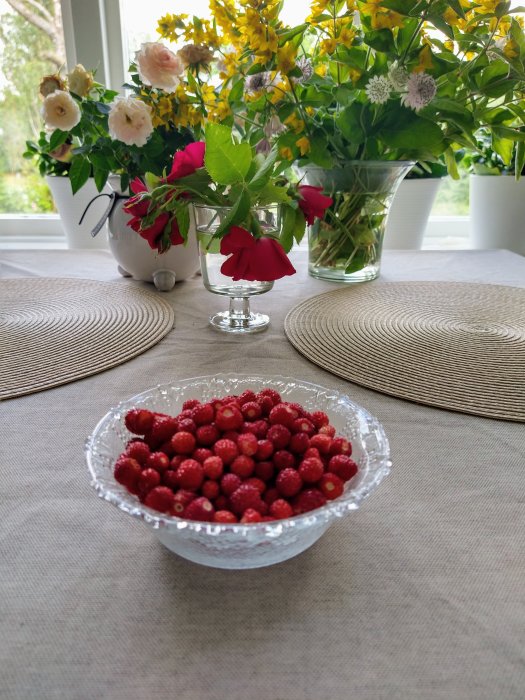 Skål med smultron framför sommarblommor och vaser på ett bord.