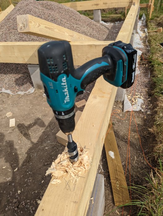 En borr uppsatt på träbjälke med borrspån, markeringslinjer syns, bakom syns en grushög.