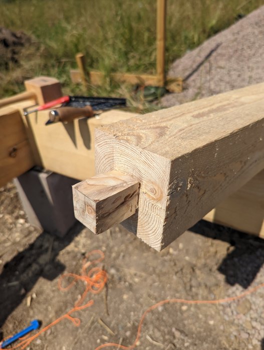 Träbjälke med uthuggen hörntapp intill ett borrat tapphål, verktyg i bakgrunden.