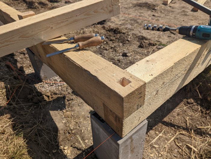 Träbjälkar med utmejslat tapphål, stämjärn, borr och borrade hål synligt på byggarbetsplats.