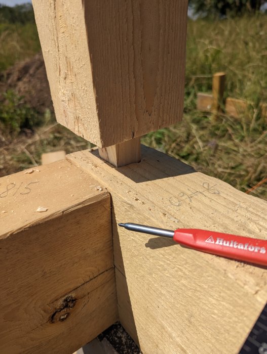 Närbild på träkonstruktion med tapp och tapphål, omgivna av verktyg och skogsbakgrund.