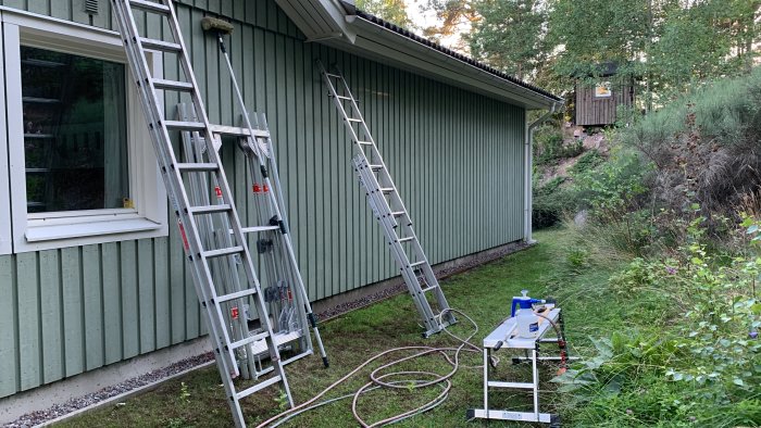 Renoveringsarbete på husfasad med stegar, tryckspruta och grönska i bakgrunden.