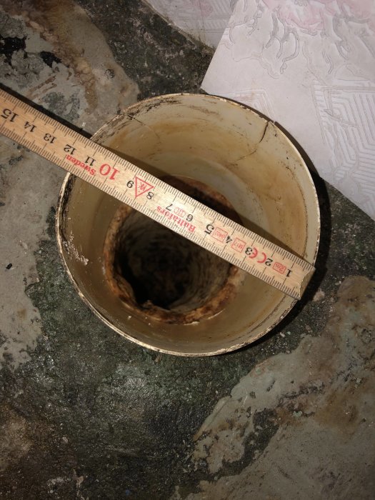 Bild av ett gammalt gjutjärnsrör i ett badrumsgolv med ett måttband över öppningen för att mäta diametern.