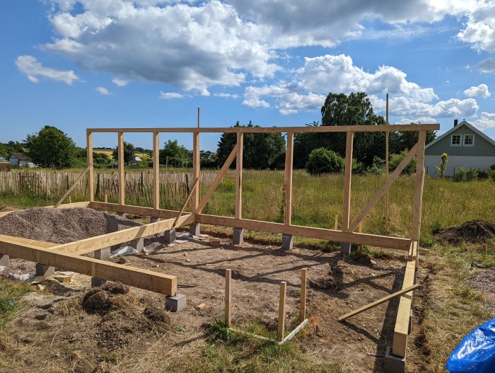 Hölje av träram och snedsträvor under konstruktion på en solig byggplats med grönska i bakgrunden.