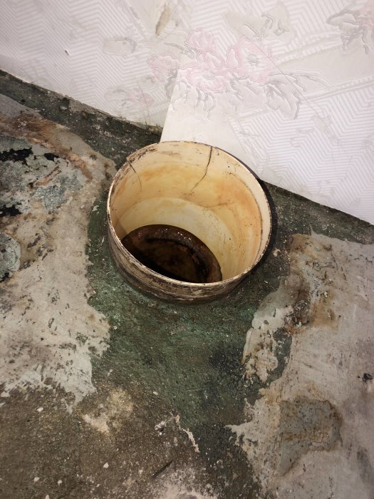 Gammalt gjutjärnsrör som sticker upp från en betonggolv med fuktskador och rester av en plastmatta.